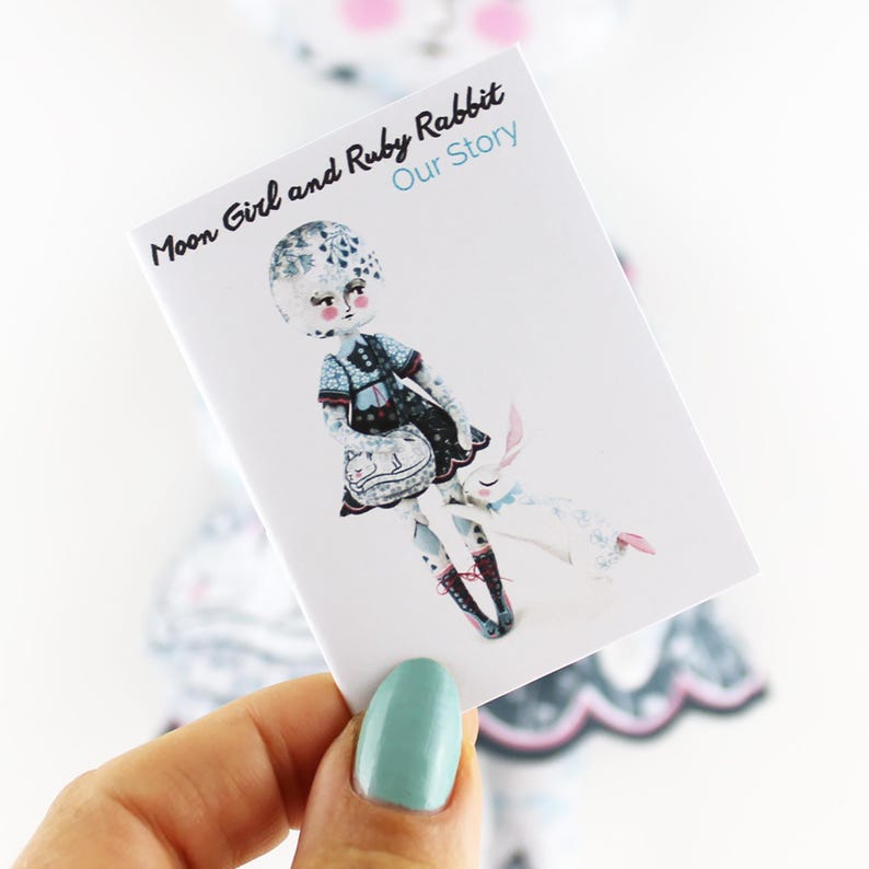 K&ouml;nnte beinhalten: Eine Karte mit einer Illustration einer Puppe und eines Kaninchens. Die Puppe tr&auml;gt ein blaues und schwarzes Kleid und tr&auml;gt eine Handtasche. Die Karte tr&auml;gt den Text "Moon Girl and Ruby Rabbit Our Story" in Schwarz und Blau.