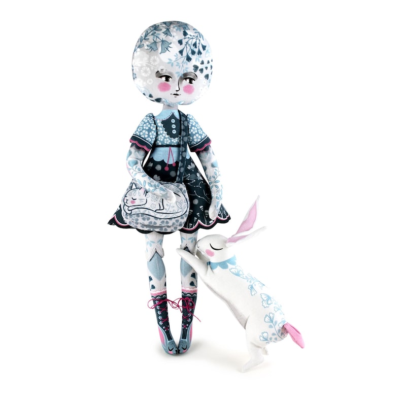 K&ouml;nnte beinhalten: Eine skurrile Puppe mit rundem Kopf und floral gemustertem Kleid, die eine Handtasche mit einem schlafenden Katzenmuster h&auml;lt. Ein wei&szlig;es Kaninchen mit rosa Ohren und floralen Akzenten steht neben der Puppe. Das Gesicht der Puppe hat rosige Wangen und einen ruhigen Ausdruck.
