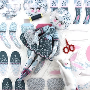 Puede incluir: Una muñeca hecha a mano con cabeza en forma de luna y vestido con estampado floral, junto con piezas de tela recortadas para una muñeca similar y un conejo blanco. La muñeca tiene mejillas rosadas y sostiene una pequeña bolsa. También se ven tijeras rojas y una bobina de hilo.