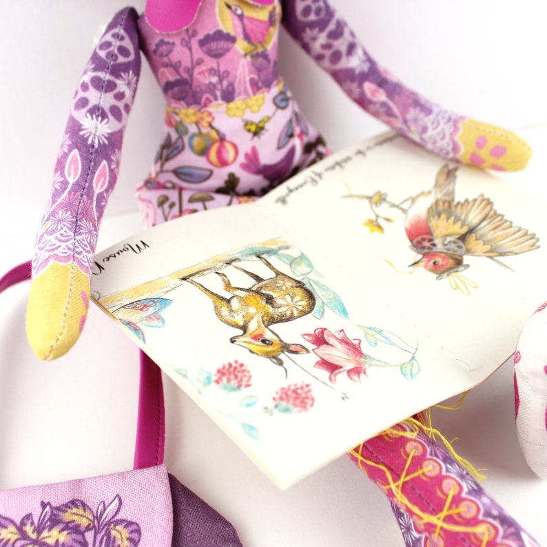 Op de afbeelding: Een pop van paarse en gele stof die een boek vasthoudt met illustraties van een hert, een vogel en bloemen. Het boek heeft de tekst "My Flower" erop.
