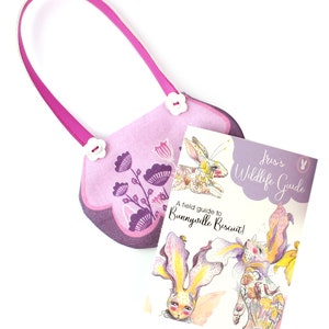 Op de afbeelding: Een roze stoffen tas met een bloemenpatroon en witte knopen. De tas is dicht en een klein boek met de titel "Iris's Wildlife Guide: A field guide to Bunnyville Biscuit!" ligt voor de tas.