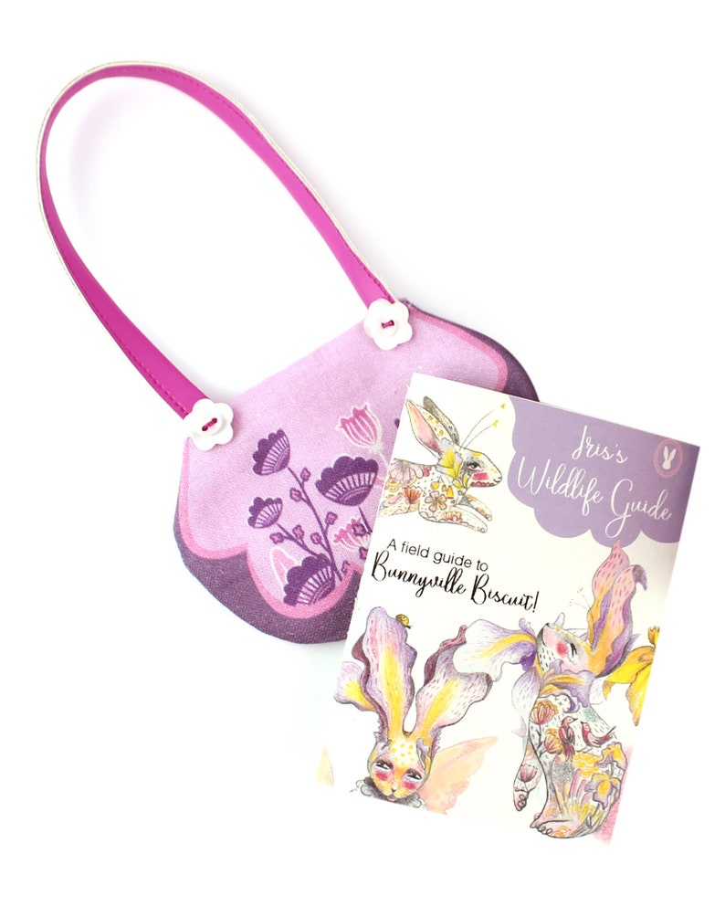 Op de afbeelding: Een roze stoffen tas met een bloemenpatroon en witte knopen. De tas is dicht en een klein boek met de titel "Iris's Wildlife Guide: A field guide to Bunnyville Biscuit!" ligt voor de tas.