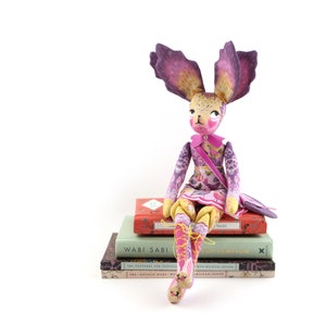 Op de afbeelding: Een grillige handgemaakte pop met paarse oren, een roze jurk en gele benen zit op een stapel boeken. De boeken hebben titels als "Wabi Sabi" en "Bed Kempion".