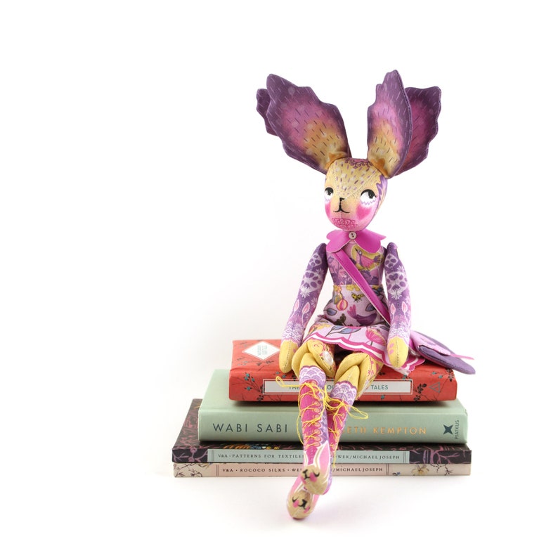 Op de afbeelding: Een grillige handgemaakte pop met paarse oren, een roze jurk en gele benen zit op een stapel boeken. De boeken hebben titels als "Wabi Sabi" en "Bed Kempion".