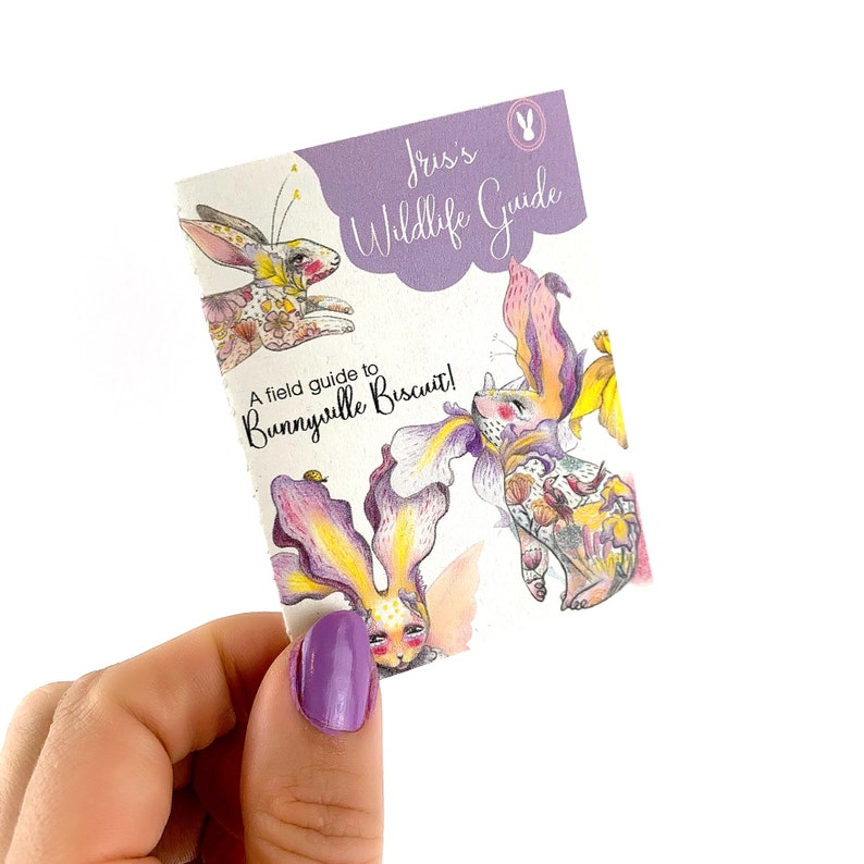 Op de afbeelding: Een ge&iuml;llustreerde gids voor Bunnyville Biscuit, met een fantasierijk konijn met een bloemenkrans en een bij. De gids is getiteld "Iris's Wildlife Guide".