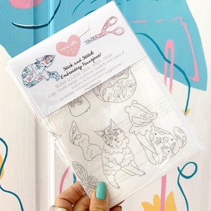 Può includere: Una confezione di plastica trasparente contenente trasferimenti per ricamo. La confezione presenta il testo "Stick and Stitch Embroidery Transfers!" e include illustrazioni di gatti, un topo, un tasso, un fungo e una farfalla. La confezione include anche le forbici.