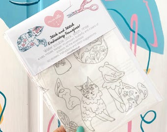 Op de afbeelding: Een doorzichtige plastic verpakking met borduurtransfers. De verpakking bevat de tekst "Stick and Stitch Embroidery Transfers!" en bevat illustraties van katten, een muis, een das, een paddenstoel en een vlinder. De verpakking bevat ook een schaar.