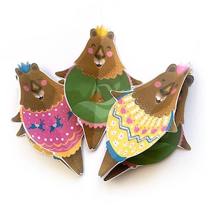 May include: Three whimsical bear-shaped paper decorations. Each bear has a brown body, a crown, and a colorful outfit. One bear wears a pink outfit with deer, another wears a green outfit, and the third wears a yellow outfit.