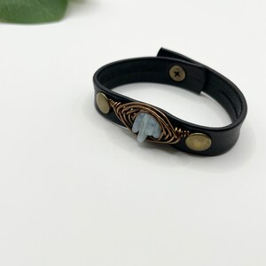 May include: A black leather bracelet with a light blue gemstone wrapped in copper wire and accented with brass accents.