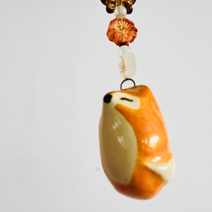May include: A hanging ornament featuring a small, smooth, orange and cream-colored fox figurine suspended from a string of decorative beads. The bead strand includes a brown butterfly-shaped bead, an orange flower-shaped bead, and clear accents.