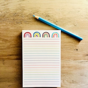 May include: A notepad with a rainbow design and lined pages, with a blue pencil resting on top. The notepad features four colorful rainbow illustrations at the top. The background is a light brown wooden surface.
