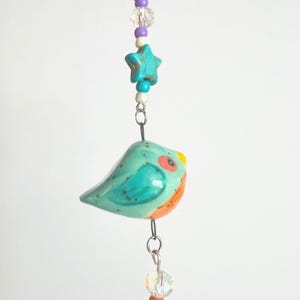 May include: A hanging ornament featuring a ceramic bird in shades of turquoise and orange with a yellow beak. The bird is suspended by a metal wire connected to a string of colorful beads, including a turquoise star-shaped bead and clear crystals.