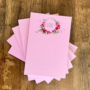 May include: A stack of six pink notepads with a floral wreath design and the word "LOVE" in the center. The wreath is made of red, pink, and purple flowers.