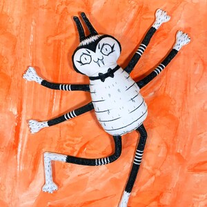 May include: A black and white spider-like plush toy with a bow tie. The toy has long, spindly legs and a large, round head with googly eyes. The toy is posed on a bright orange background.