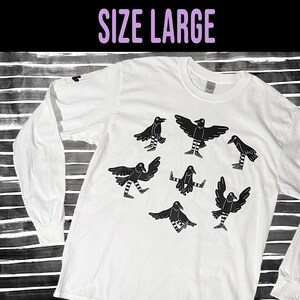 May include: White long-sleeved t-shirt with a black graphic of seven crows with striped legs. The text "SIZE LARGE" is printed in purple at the top of the image.