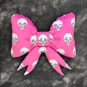 Peut inclure: Un nœud en peluche rose avec un motif de crâne. Le nœud est fait d'un tissu doux et duveteux et a un contour blanc autour de chaque crâne.