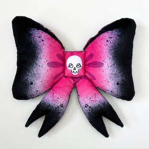 Peut inclure: Un nœud rose et noir avec un motif de crâne blanc au centre. Le nœud est fait d'un tissu doux et a un motif moucheté.