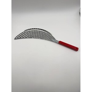 May include: A crescent-shaped metal colander with a perforated surface featuring numerous circular holes. The kitchen tool includes a long, straight red handle attached to the curved metal body with a visible silver metal rivet.