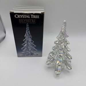 May include: A clear, iridescent glass Christmas tree figurine with a tiered, spiraled design. Beside it stands a black rectangular box labeled "CRYSTAL TREE" and "SILVESTRI THE WONDER OF CHRISTMAS" with a graphic of the same tree on the front.