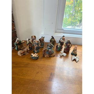 May include: A collection of fifteen small, hand-painted ceramic nativity figurines including figures of people, an angel, a camel, a donkey, a sheep, and a separate pair of white wings, all arranged on a smooth wooden surface.