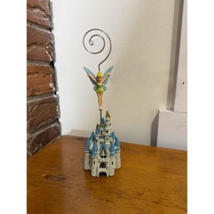 May include: A decorative figurine featuring a miniature castle with blue turrets, topped with a fairy figure and a silver wire spiral. The fairy has green wings and a yellow dress. The castle is beige and white.