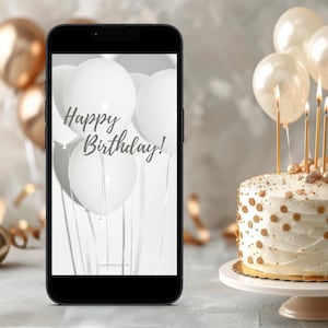 Könnte beinhalten: Ein Smartphone zeigt einen weißen Hintergrund mit weißen Ballons und dem Text "Happy Birthday!" Im Hintergrund ist ein weißer Kuchen mit goldenen Streuseln und Kerzen zu sehen.