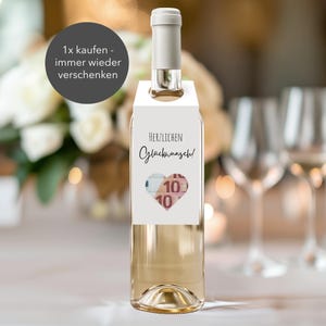 Könnte beinhalten: Eine klare Weinflasche mit einem weißen Etikett mit dem Text "Herzlichen Glückwunsch!" und einem herzförmigen Design mit einem 10-Euro-Schein. Die Flasche hat einen weißen Hals und steht auf einem Tisch mit Blumen und Gläsern im Hintergrund.