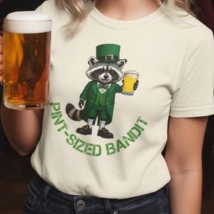 May include: Cream-colored t-shirt featuring a cartoon raccoon in a green suit and top hat, holding a beer. The text "PINT-SIZED BANDIT" is printed in green. A woman is holding a beer mug.