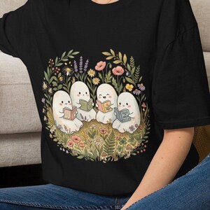 May include: A black t-shirt featuring a graphic print of four white ghosts sitting in a circle in a field of colorful wildflowers, each ghost holding and reading a small book, with one book clearly labeled with the text Tales of Magic.
