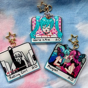 May include: Three acrylic keychains with gold star clasps. Each keychain features a different anime-style illustration. One says "World is Mine", another says "Rolling Girl", and the last says "Magnet". The background is a tie-dye pattern.