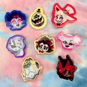 May include: Eight colorful cartoon character pins featuring characters from the animated series "Hazbin Hotel". The pins are made of clear acrylic and feature a variety of characters, including a red demon with horns, a white angel with wings, and a yellow pumpkin-like creature.