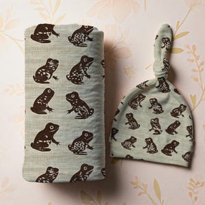 May include: A baby blanket and knotted hat set. The light blue fabric is patterned with brown frog illustrations. The blanket is rectangular and folded. The hat is a light blue color with a knotted top.
