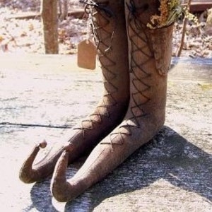 Könnte beinhalten: Ein Paar braune Stoffstiefel mit aufgebogenen Spitzen, schwarzer Schnürung und beigefarbenen, gemusterten Stoffoberteilen, die als Nadelkissen dienen. Jeder Stiefel ist mit einer farbigen Nadel besteckt und trägt ein kleines, leeres braunes Papieretikett.