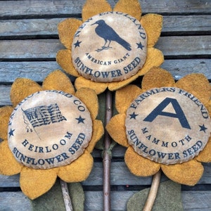 May include: Three handcrafted sunflower decorations with tan centers and yellow petals. Each center features a vintage-style design with text and illustrations, including a crow, an American flag, and the letter "A". The stems are made of natural twigs.