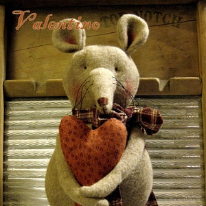 May include: A handmade, plush mouse figurine holding a heart. The mouse is gray with a plaid scarf and a heart-shaped pillow. The text "Valentino" and "Sassafras Hill Primitives" are visible. The figurine is set against a rustic wooden background.