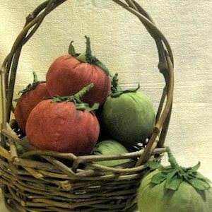 May include: A wicker basket filled with red and green fabric tomatoes. The basket is made of woven twigs and has a handle. The tomatoes are arranged in a pile, with some spilling out of the basket. The text "Olde Farmstead Tomatoes" is written in red at the top of the image. The text "Sassafras Hill Primitives" is written in red at the bottom of the image.