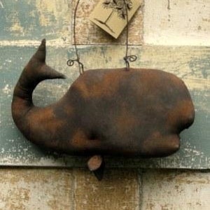 Könnte beinhalten: Ein braunes, walförmiges Stoffornament mit einer rostigen braunen Oberfläche. Der Wal hängt an einem Drahtbügel und hat ein Etikett mit der Aufschrift "Rusty Olde Whale" und "Sassafras Hill Primitives 285-09".