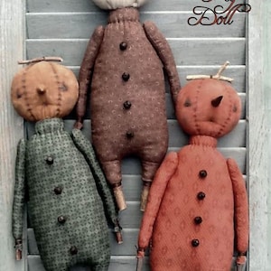 May include: Three fabric dolls with round pumpkin-shaped heads and twig stems. The dolls wear brown, green, and orange patterned outfits with dark buttons down the front. Text on the image reads Pumpkin Patch Peg Doll and Sassafras Hill Primitives.