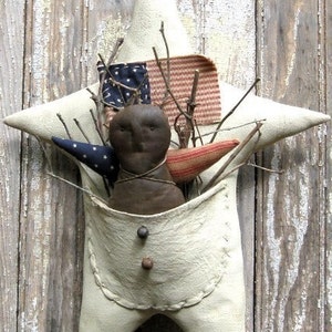 Könnte beinhalten: Cremefarbene sternförmige Tasche mit einer primitiven Puppe im Inneren. Die Tasche zeigt eine kleine amerikanische Flagge und Zweige. Die Puppe hat einen dunkelbraunen Kopf und ist mit Bindfaden gebunden. Der Stern ist auf einem verwitterten Holzgrund befestigt.