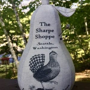 Könnte beinhalten: Birnenförmiges Nadelkissen aus hellgrauem Stoff mit einer Hahn-Illustration und dem Text "The Sharpe Shoppe, Seattle, Washington" und "Haystack Needle Makers". Das Nadelkissen hat eine Schlaufe und ein Blattdetail.