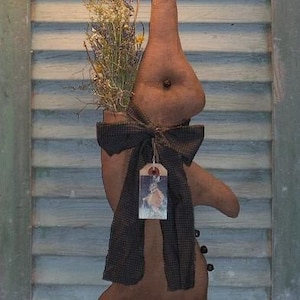 Könnte beinhalten: Ein brauner, stoffbezogener Hasenaufhänger mit langer Nase und Fliege. Er hält Trockenblumen und hat ein Etikett mit einem Hasenbild. Der Text "Gathering Rabbit Hanger" und "Sassafras Hill Primitives 08" sind sichtbar.