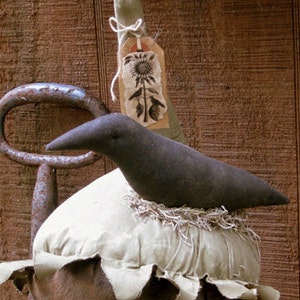 Könnte beinhalten: Ein handgefertigter, rustikaler Zier-Vogel, der auf einer nestartigen Struktur sitzt. Der Vogel ist schwarz, und das Nest besteht aus einem hellfarbigen Stoff mit einem braunen, verwitterten Sockel. Ein Anhänger mit einer Sonnenblumen-Illustration hängt darüber.