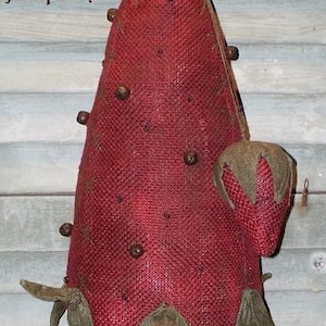 May include: A handcrafted, red burlap strawberry-shaped pin cushion with brown accents. The pin cushion has a textured surface and is adorned with small, round, brown embellishments. The base is a light brown color, and the text "Berry Olde Pinpoke" is visible.
