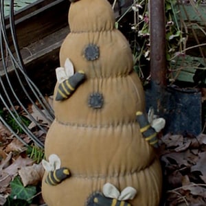Könnte beinhalten: Ein dekoratives, beige-farbenes Bienenhaus-Ornament mit mehreren Stoffbienen. Die Bienen haben gelb-schwarze Streifen und weiße Flügel. Das Ornament hat ein genähtes Design und kleine graue Flecken. Der Text "Skep O Bumbles" und "Sassafras Hill Primitives" sind sichtbar.