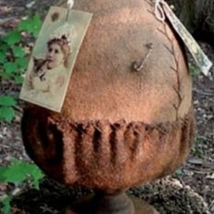 Könnte beinhalten: Ein handgefertigtes, eichelförmiges Dekorationsstück. Es besteht aus braunem Stoff und steht auf einem Holzsockel. Ein Bild im Vintage-Stil ist vorne angebracht. Die Oberseite der Eichel hat ein kleines, dekoratives Element. Für die Inneneinrichtung.