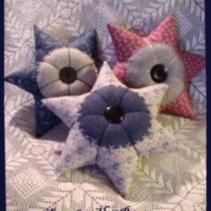 Könnte beinhalten: Drei sternförmige Nadelkissen in Blau- und Rosatönen, mit einem zentralen kreisförmigen Design und einem Knopf. Die Kissen sind mit floralen Mustern und dem Text "Six Point Pincushion Pillows" und "Sassafras Hill Primitives" verziert.