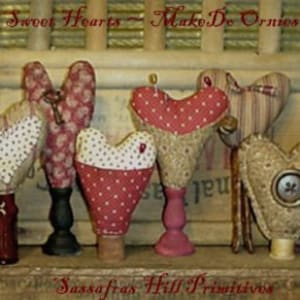 Könnte beinhalten: Eine Sammlung handgefertigter Stoffherz-Ornamente in verschiedenen Mustern und Farben, darunter rote Punkte und Streifen. Jedes Herz ist auf einem Holzsockel befestigt, mit dem Text "Sweet Hearts - MakeDo Ornies" und "Sassafras Hill Primitives".