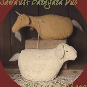 Op de afbeelding: Een wit schaap en een bruine geit, beide van stof, zitten op een houten oppervlak. Het schaap is links en de geit rechts. De geit heeft een lange staart en het schaap een korte staart. De geit kijkt naar links en het schaap naar rechts. De tekst "Sawdust Banyard Duo" staat bovenaan de afbeelding en de tekst "Billygoat & Sheep" onderaan de afbeelding.