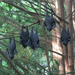 May include: Five black felt bat decorations hanging from tree branches in a forest setting.
