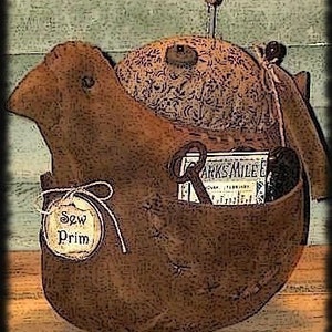 Könnte beinhalten: Eine braune, vogelartige Nähbox mit einem Nadelkissen, Garnrollen und einer kleinen Stoffbox mit dem Text "Sew Prim" darauf. Die Box ist mit Nähten verziert und hat einen rustikalen, Vintage-Look. Der Text "Early Sewing Bird" befindet sich oben im Bild und "Sassafras Hill Primitives 266-06" unten.